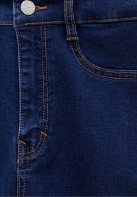Gros plan sur un jean en denim bleu foncé montrant la poche avant, la passants de ceinture, les coutures orange et un bouton argenté gravé "Pull&Bear".