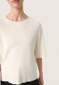 White short-sleeve top with a round neckline, made of textured fabric, features a slightly scalloped hem. Simple and minimalist design.