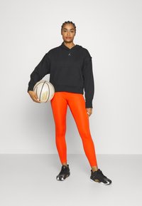 Felpa nera con cappuccio e tasca frontale, leggings arancioni e scarpe da basket nere. Modella che tiene una palla da basket, con capelli intrecciati.