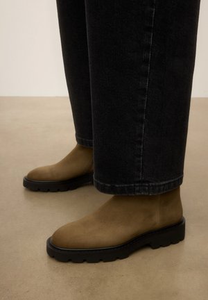 Personne portant des bottes en daim marron avec des semelles épaisses noires et un pantalon en denim noir debout sur un sol beige.