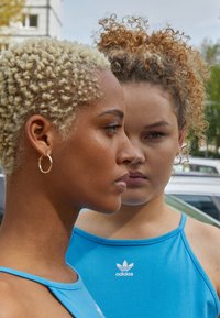 Dvi moterys su skirtingomis šukuosenomis, viena su trumpais blonde garbanais, o kita su ilgais garbanais, abi dėvi mėlynas sportines marškinėles su Adidas logotipu.