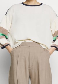 Krämfärgad tröja med ribbade, flerfärgade mönstrade ärmar i grönt, brunt och blått. Kombinerad med ljusbeiga högmidjade byxor.