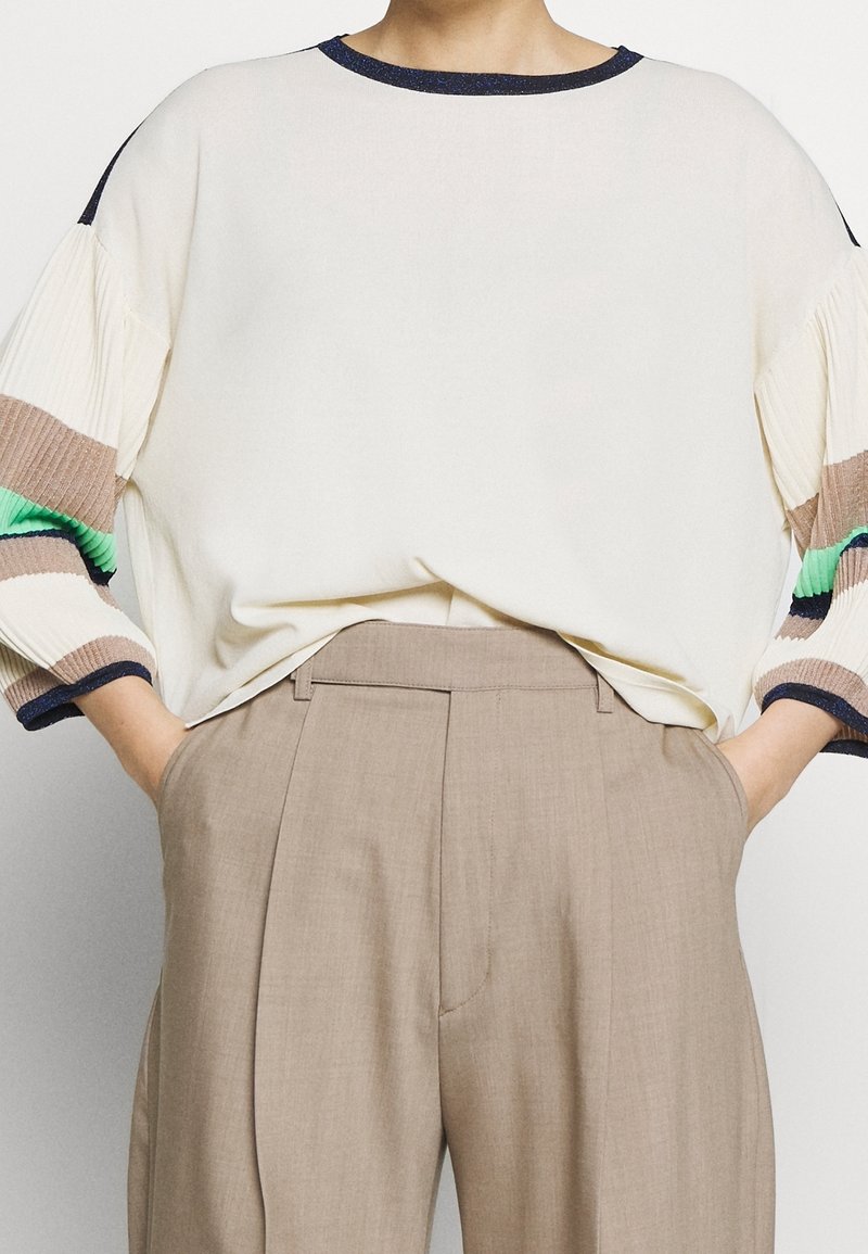 Krämfärgad tröja med ribbade, flerfärgade mönstrade ärmar i grönt, brunt och blått. Kombinerad med ljusbeiga högmidjade byxor.