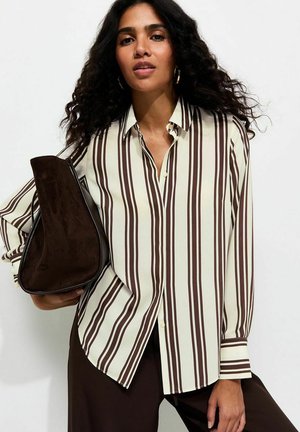 Vrouw met lang krullend haar die een crèmekleurige blouse draagt met bruine verticale strepen en donkerbruine broek, en een donkerbruine suède handtas vasthoudt.