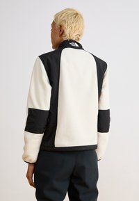 Čierna a smotanová fleece bunda s vysokým golierom, ktorá má čierne nylonové akcenty a kontrastný patchwork dizajn na rukávoch a chrbte.