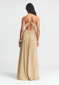 Maxi sukienka z beżowego materiału, o luźnym kroju, z krzyżowanym dekoltem z tyłu i otwartym plecami. Podkreślona subtelną teksturą i swobodnym ruchem.