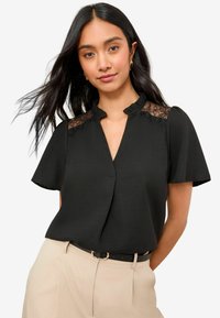 Blouse noire avec un empiècement en dentelle, manches courtes et col mandarin, associée à un pantalon beige taille haute et une ceinture noire.