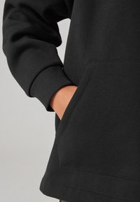 Sudadera negra con bolsillo tipo canguro; tejido texturizado; puños acanalados; corte relajado; mostrando la mano en el bolsillo y detalle del dobladillo.