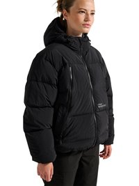 Kobieta w czarnej pikowanej kurtce z kapturem, zamkiem z przodu i kieszeniami na zamek, stojąca na białym tle.