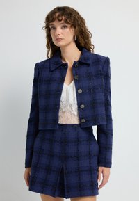 Marinblå tweedkortjacka med rutmönster, som har metalliska accenter, fem knappar och krage. I kombination med en matchande kjol.