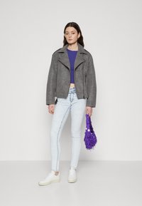 Veste de moto en daim gris, crop top violet, jeans skinny bleu clair, baskets blanches, tenant un sac à main texturé violet avec des accents.