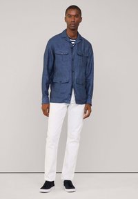 Giacca di jeans blu scuro con una finitura testurizzata, caratterizzata da quattro tasche frontali, chiusura a bottoni e colletto. Indossata sopra una camicia a righe.