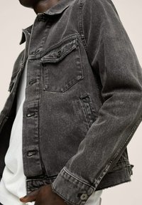 Uomo che indossa una giacca di denim nera sbiadita sopra una camicia bianca, con una mano appoggiata vicino alla vita, su uno sfondo chiaro e neutro.