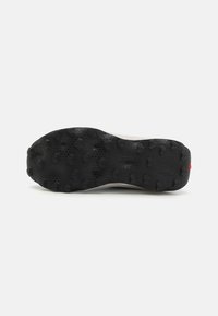 Semelle de chaussure en caoutchouc noir avec des crampons multidirectionnels, surface texturée et légère courbure. Présente un accent rouge au niveau du talon.
