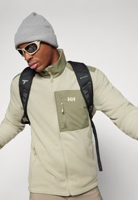 Chaqueta de forro polar en color beige claro con un bolsillo con cremallera en verde oliva, combinada con un gorro de lana gris y gafas de sol. Se ven las correas de la mochila.