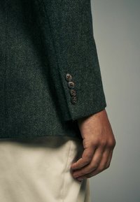 Dunkelgrüner Hahnentrittmantel mit strukturiertem Finish und vier braunen Knöpfen an den Ärmeln, kombiniert mit hellbeigen Hosen.