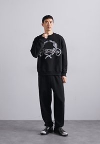 Mann trägt einen schwarzen Moschino-Pullover und lockere schwarze Hose, steht mit einer Hand nahe seinem Hals vor einem schlichten hellen Hintergrund.