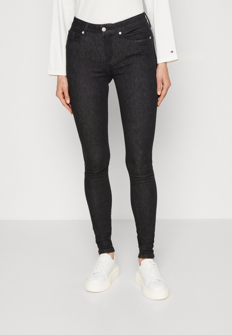 Person trägt schwarze Skinny-Jeans, weißes Langarmshirt und weiße Plateau-Sneaker und steht vor einem schlichten Hintergrund.