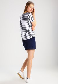 Gestreepte korte mouwen top in wit en marineblauw, gecombineerd met een marineblauwe spijkerrok. Witte sneakers met een metalen accent. Model staat met de rug naar de camera.