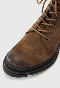Brown suede ankle boot with round toe, metal eyelets, and matching laces. Features a textured black rubber sole with tread for grip.