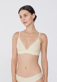 Béžový bezdrátový bralet s hlubokým výstřihem do V, nastavitelné ramínka a bezešvý design; měkká textura a minimalistický styl.
