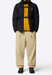 Chaqueta tipo plumón negra con puños elásticos, superpuesta a un forro polar amarillo, combinada con pantalones beige holgados que tienen bolsillos con cremallera.