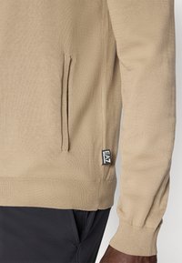 Μπεζ πλεκτό φούτερ με πλαϊνή τσέπη, ριμπ μανσέτες και ετικέτα με λογότυπο. Το ύφασμα έχει ομαλή, υφή με ανάγλυφη όψη.