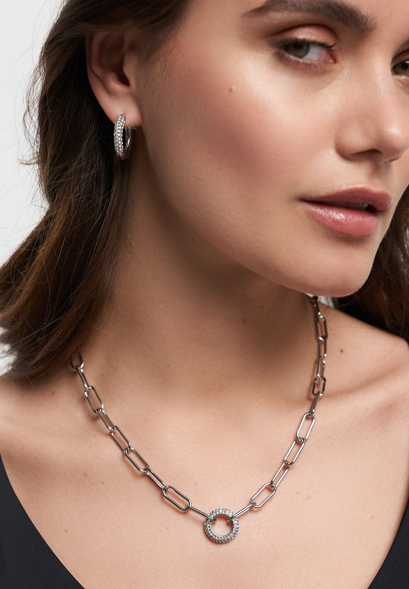 Silberne Metallkette mit einem kreisförmigen Anhänger, der mit kleinen Strasssteinen besetzt ist. Passende Creolen mit Strassakzenten.