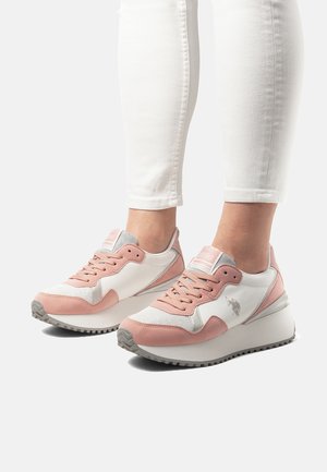 Białe i różowe sneakersy na platformie noszone z białymi dopasowanymi spodniami, pokazujące widok z przodu i z boku z widocznymi sznurowadłami i detalem z logo.