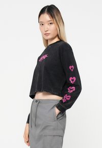 Cămașă neagră, scurtă, cu mâneci lungi, cu modele grafice roz pe față și mâneci. Fabricată dintr-un material moale, texturat. Asortată cu pantaloni cargo gri.