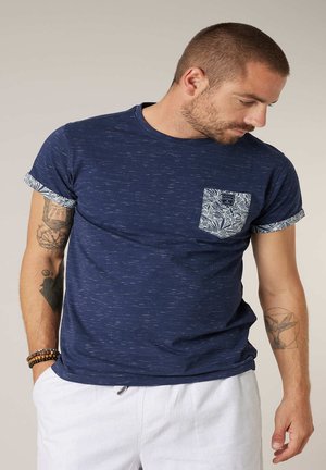 Homme portant un T-shirt bleu marine avec une poche à motif et des manches retroussées, un pantalon blanc et des bracelets, regardant vers le bas à sa droite.