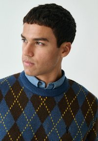 Strickpullover mit einem Rautenmuster in Braun, Blau und Gelb, einem blauen Rippenkragen sowie einem darunter getragenen Jeanshemd mit Kragen.