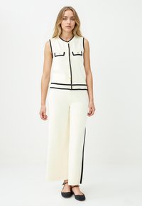 Mouwloze crèmekleurige top met zwarte accenten en knopen, gecombineerd met crèmekleurige wijde broek met zwarte zijstrepen. Gladde textuur.