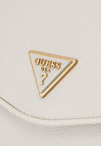 Materiale bianco texturizzato con un logo triangolare oro e bianco che presenta "GUESS USA" e un dettaglio a punto interrogativo.