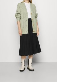 Světlý zelený pletený cardigan s žebrovanou strukturou, na tři knoflíky, se dvěma kapsami, nošený přes bíl ý tričko a černou skládací sukni; bílé kotníkové boty.