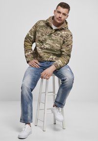 Kamuflage pullover jakke i nuancer af grøn og brun, med en frontlomme, parret med lyseblå jeans og hvide sneakers.