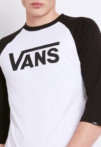Hvid baseballstil t-shirt med sorte raglanærmer; prydet med en fremtrædende sort "VANS" logo på brystet. Blødt bomuldsstof.