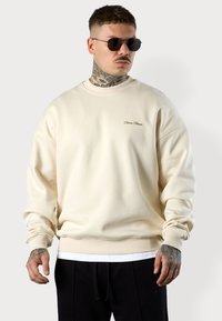Sweatshirt oversize couleur crème avec des poignets et un col côtelés, arborant une petite broderie de logo sur la poitrine. Tissu doux et texturé.