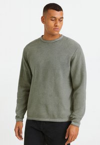 Grau gestreift, langärmliger Sweatshirt mit Rundhalsausschnitt. Strukturierten Stoff, lässiger Schnitt. Getragen mit schwarzen Hosen, Hände entspannt an den Seiten.