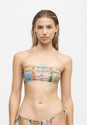 Jeune femme aux cheveux mouillés portant un bikini bandeau multicolore à motif coquillages et un collier blanc en coquillages sur fond blanc.