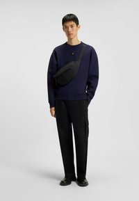 Ung man som bär en marinblå sweatshirt, svarta cargobyxor, svarta skor och en svart axelväska mot en enkel grå bakgrund.