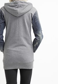 Felpa grigia con maniche in denim, caratterizzata da un cappuccio con lacci, polsini a costine e una vestibilità lunga e comoda. Il denim vissuto aggiunge delle texture.