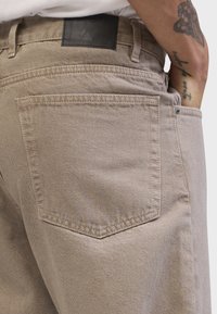 Pantalones de mezclilla beige con cintura de altura media, que cuentan con un parche de cuero negro en la parte posterior, un bolsillo trasero redondeado y una textura sutil.