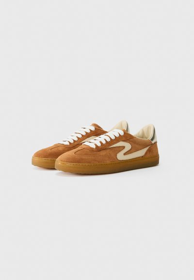 Sneaker in suede marrone con lacci bianchi, un motivo laterale beige e una suola in gomma naturale. Texture morbida e forma arrotondata.