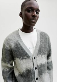 Šedý a bílý melírovaný cardigan s výstřihem do V, zdobený černými knoflíky a měkkou, texturovanou látkou, nošený přes bílou košili s vyšitým textem.