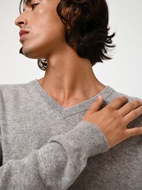 Grauer Strickpullover mit V-Ausschnitt, ausgestattet mit gerippten Bündchen und einer glatten Textur. Das Model berührt die Schulter und zeigt die Passform des Kleidungsstücks.