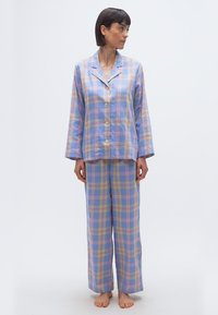 Γυναίκα όρθια και ξυπόλητη, φορώντας πουκάμισο πιτζάμας με παστέλ καρό μοτίβο και κουμπιά, με μακριά μανίκια, και ταιριαστό παντελόνι με φαρδιά πόδια, κοιτάζοντας προς τα αριστερά της.