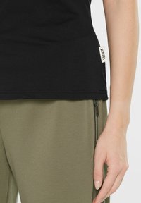 Schwarzes Baumwoll-T-Shirt mit geradem Saum und einem kleinen Stoffetikett an der Seite. Kombiniert mit olivgrünen, strukturierten Hosen mit Reißverschluss.