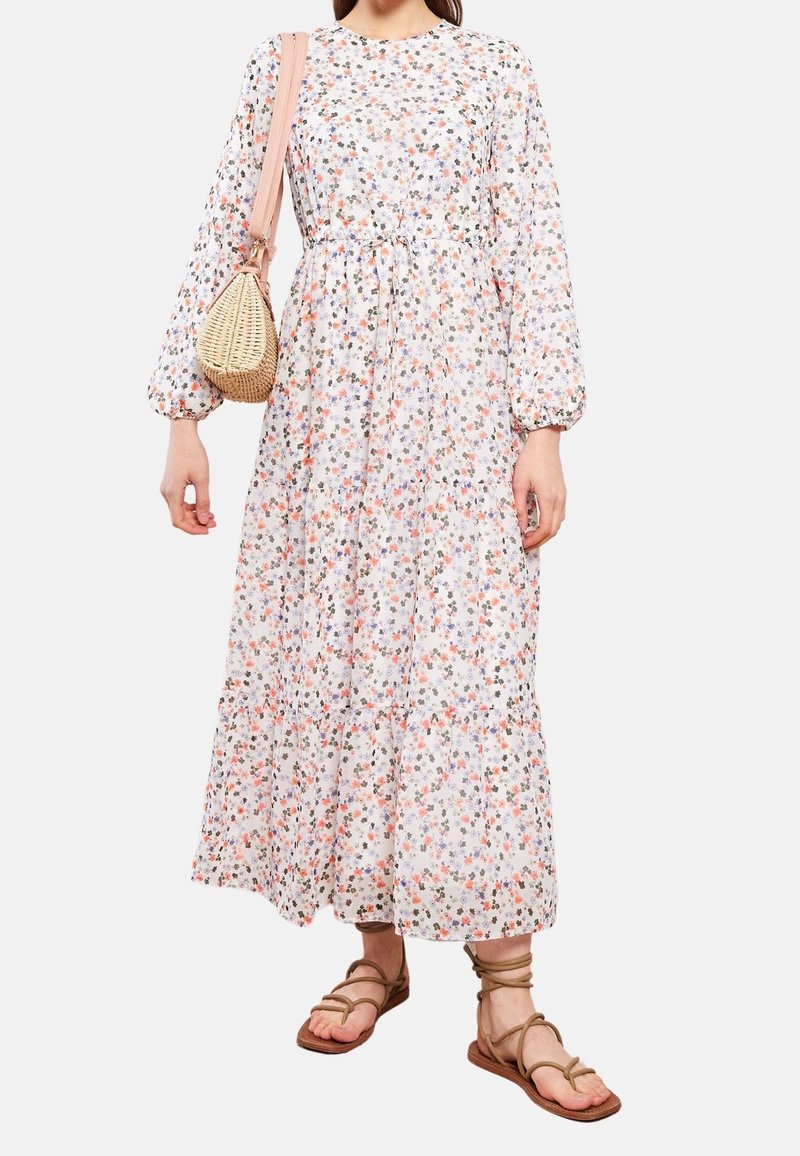 Robe maxi à manches longues, florale, en blanc avec des motifs roses, bleus et verts. Elle possède une taille cintrée et une jupe à volants ; accompagnée d'un sac tissé.