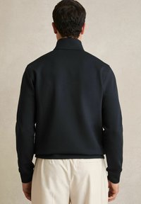 Hanorac negru cu guler înalt, textură netedă, design mulat și mâneci lungi, asortat cu pantaloni deschisi la culoare.